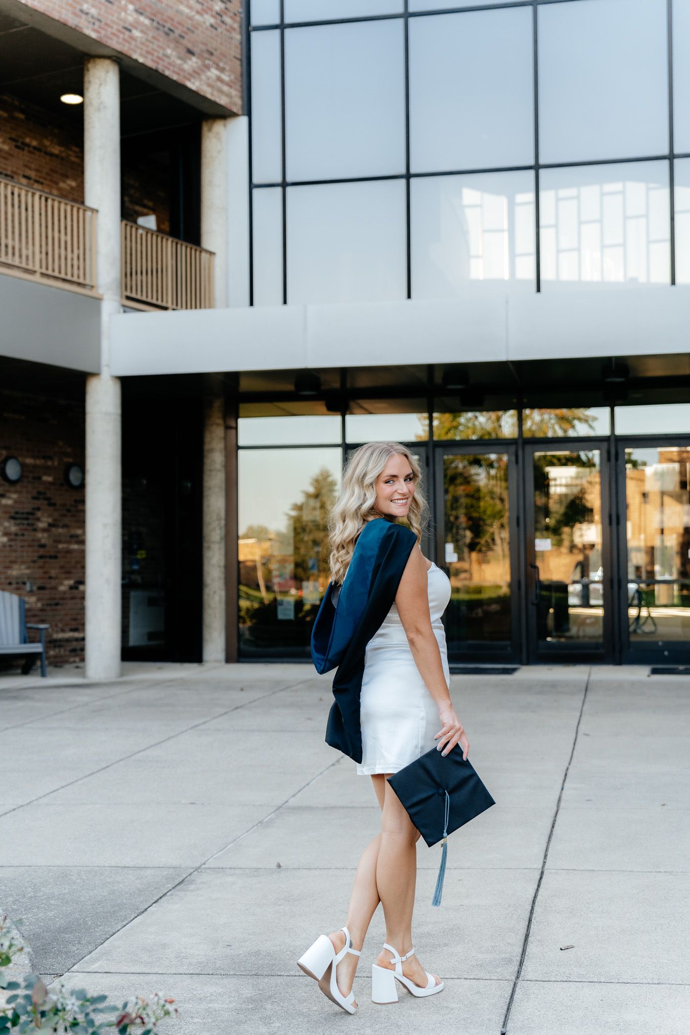 Graduate in white dress and navy stole poses confidently outside modern building.