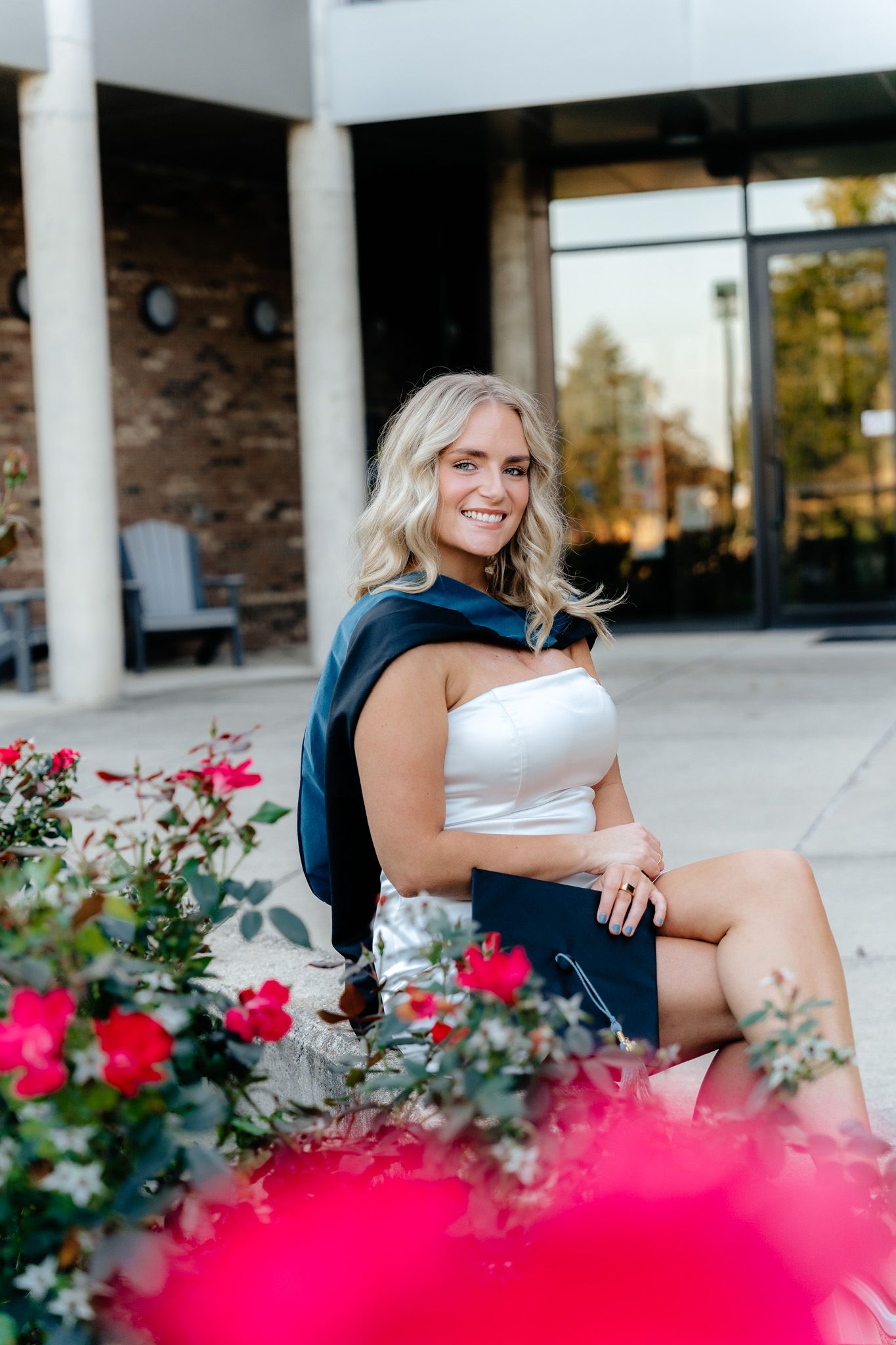 Graduate in white dress sits near blooming flowers with navy graduation stole.