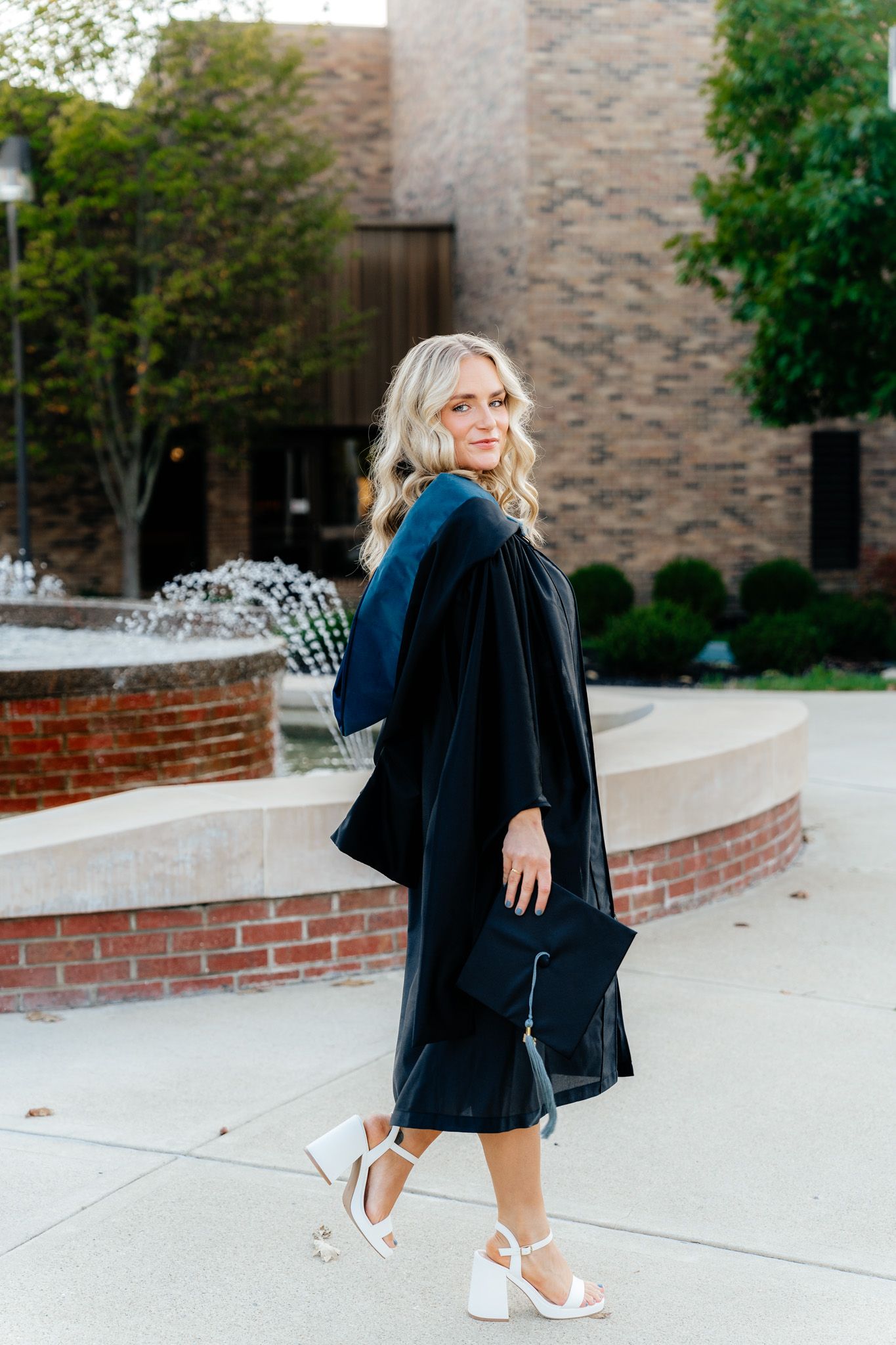 Graduate in academic regalia walks past fountain on campus grounds.