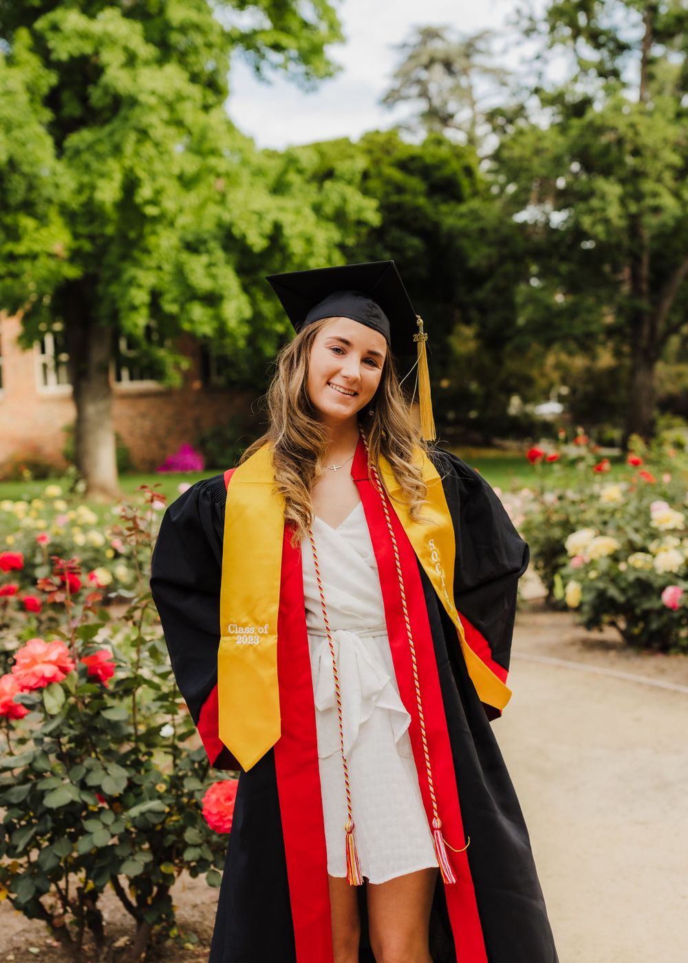 Graduate in black cap and gown with gold and red stoles posing in a rose garden on graduation day.