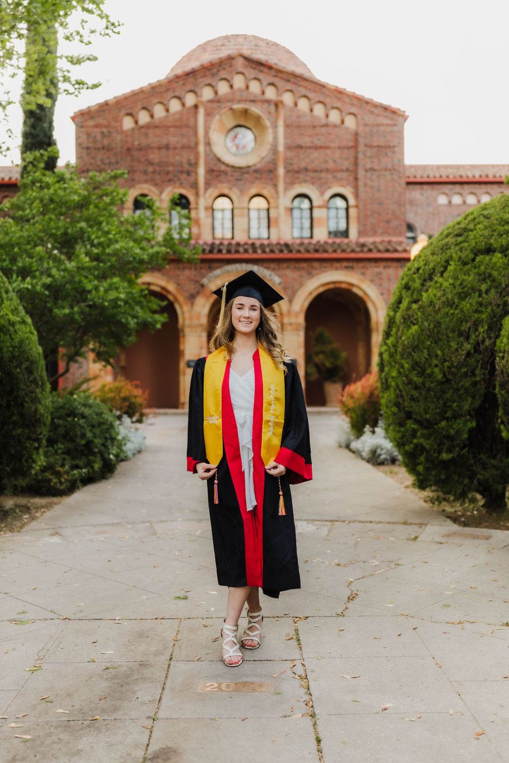 Graduate in cap and gown standing before historic Spanish mission-style building with arched colonnade and terracotta roof.