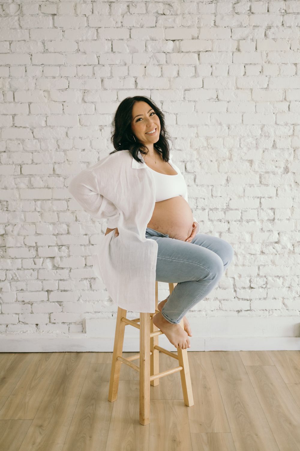 An expectant mother poses on a wooden stool against a white brick wall.