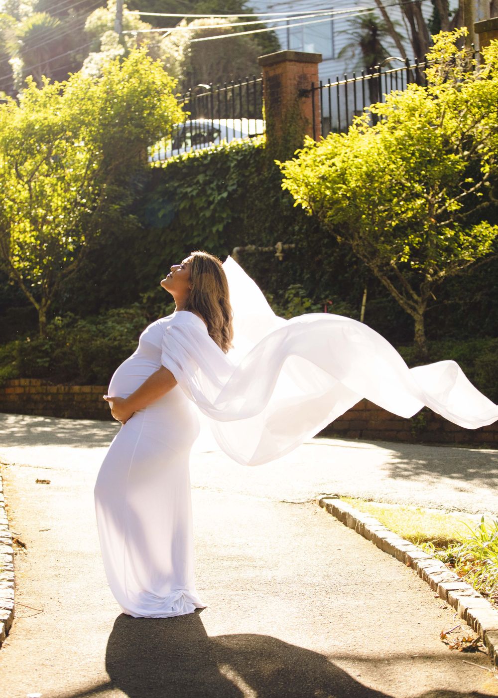 Outdoor maternity portrait of backlit pregnant woman in a white dress with floating white tulle.