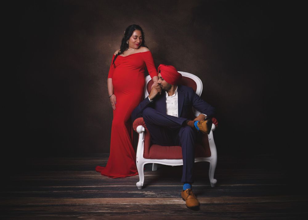 Portrait of couple with pregnant woman standing in red dress while her husband is sitting and kissing her hand.