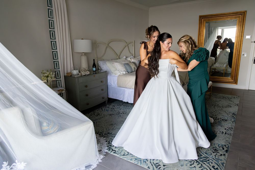 A bride in a white ball gown wedding dress gets ready in a sunlit room with bridesmaids in green dresses.