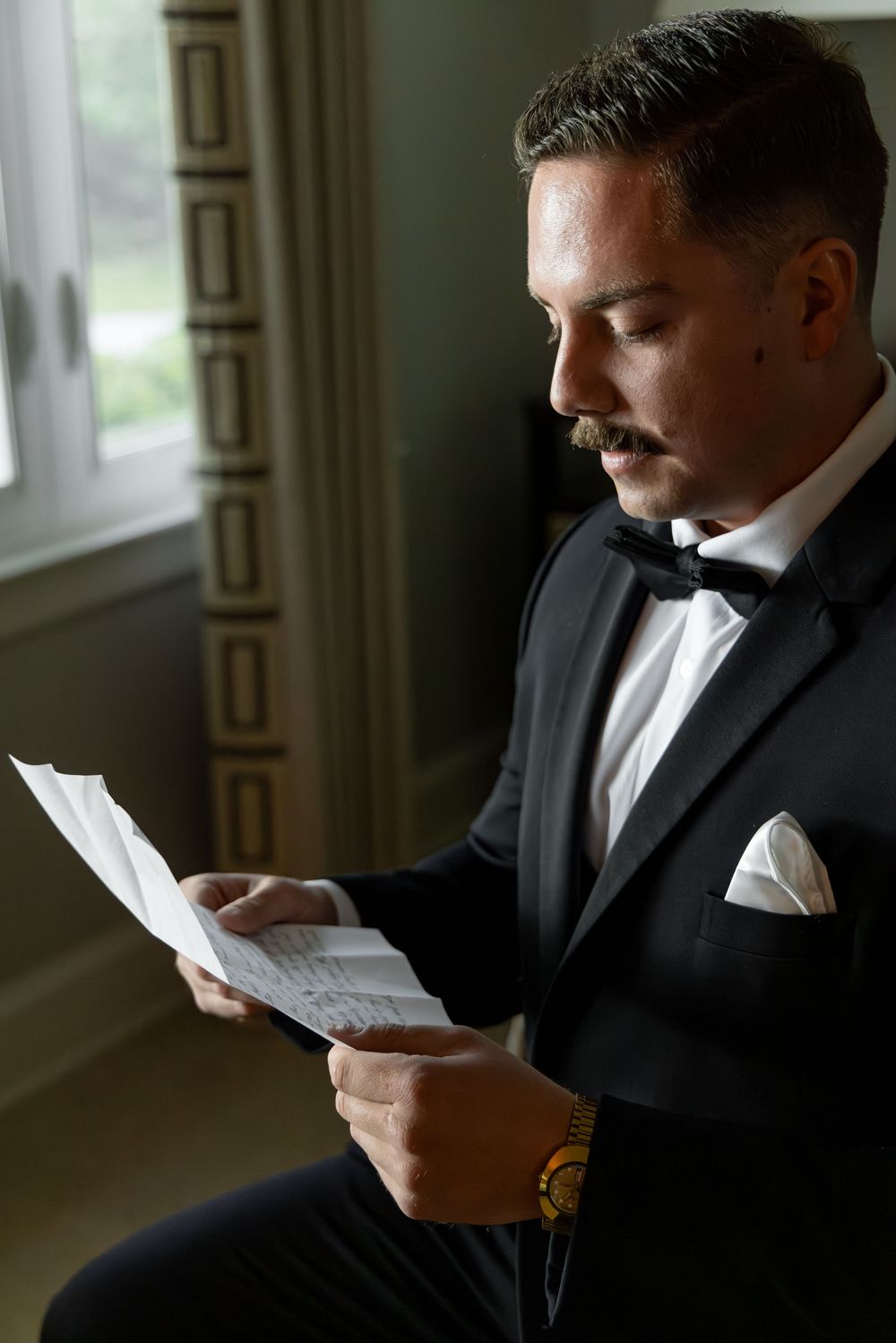 Groom in black tuxedo reads emotional letter in moody window lighting.