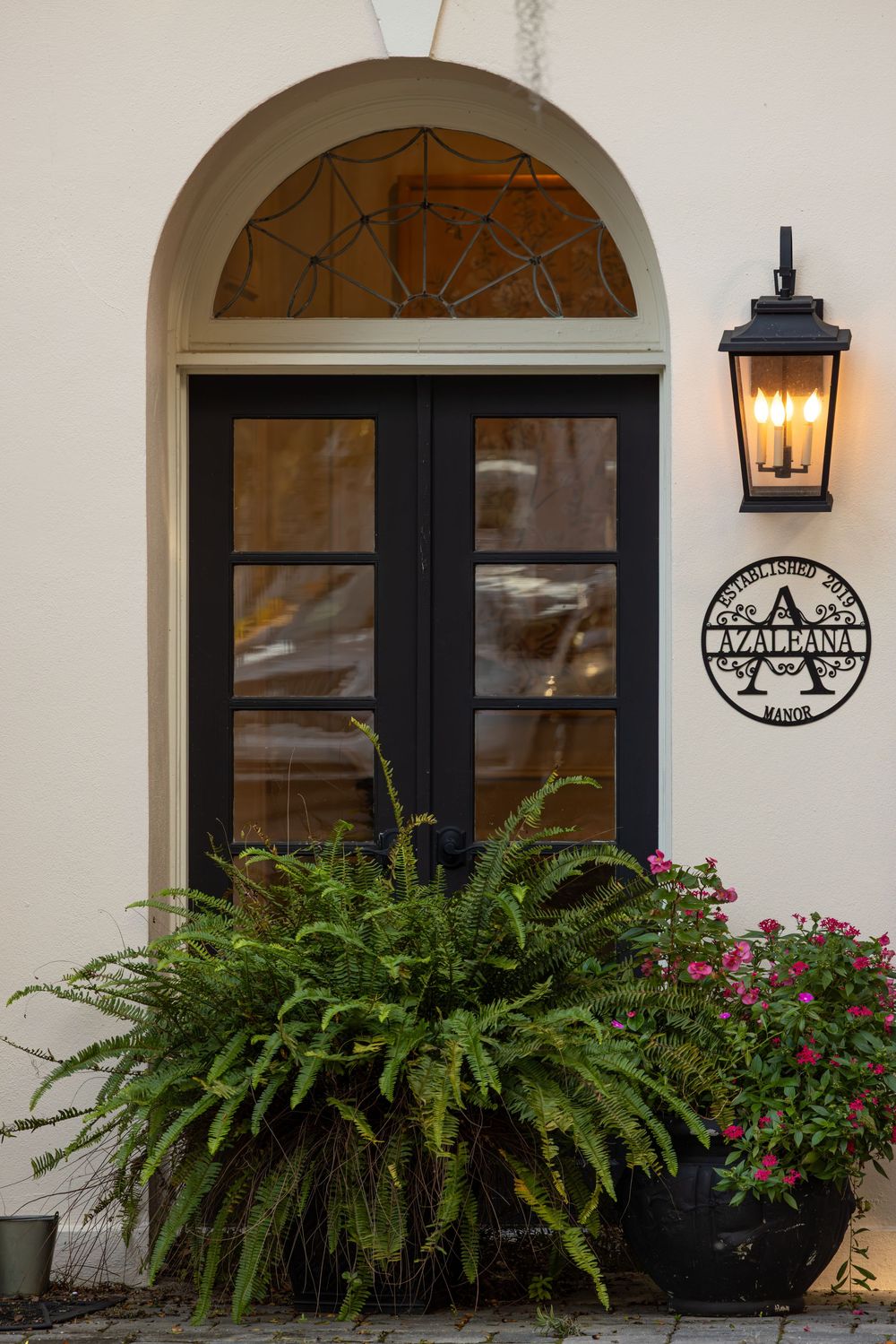 Elegant entrance door with arched transom, lantern light fixture and lush fern planter below.