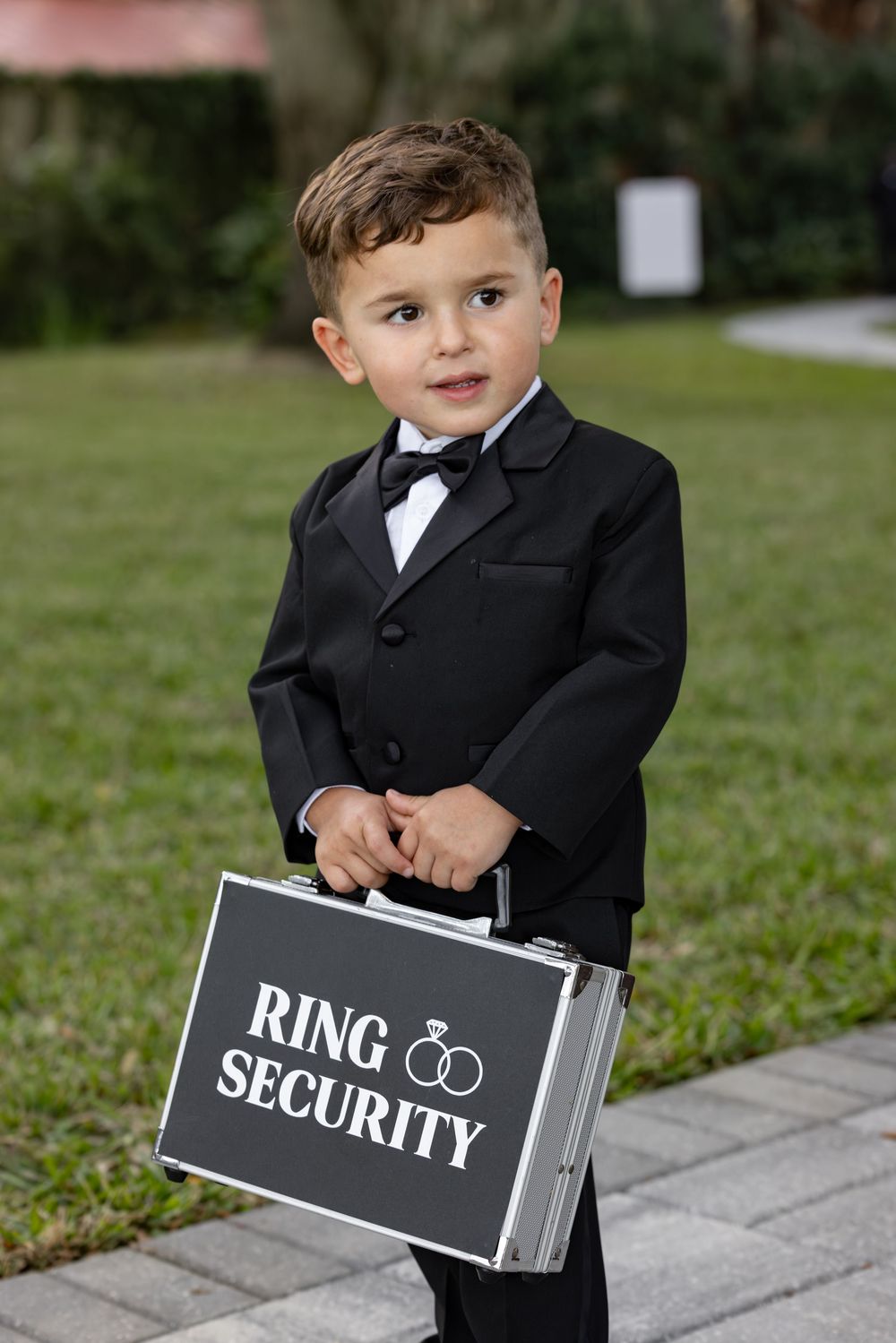Young ring bearer in a black suit carrying a silver briefcase labeled 'Ring Security' on a garden path.