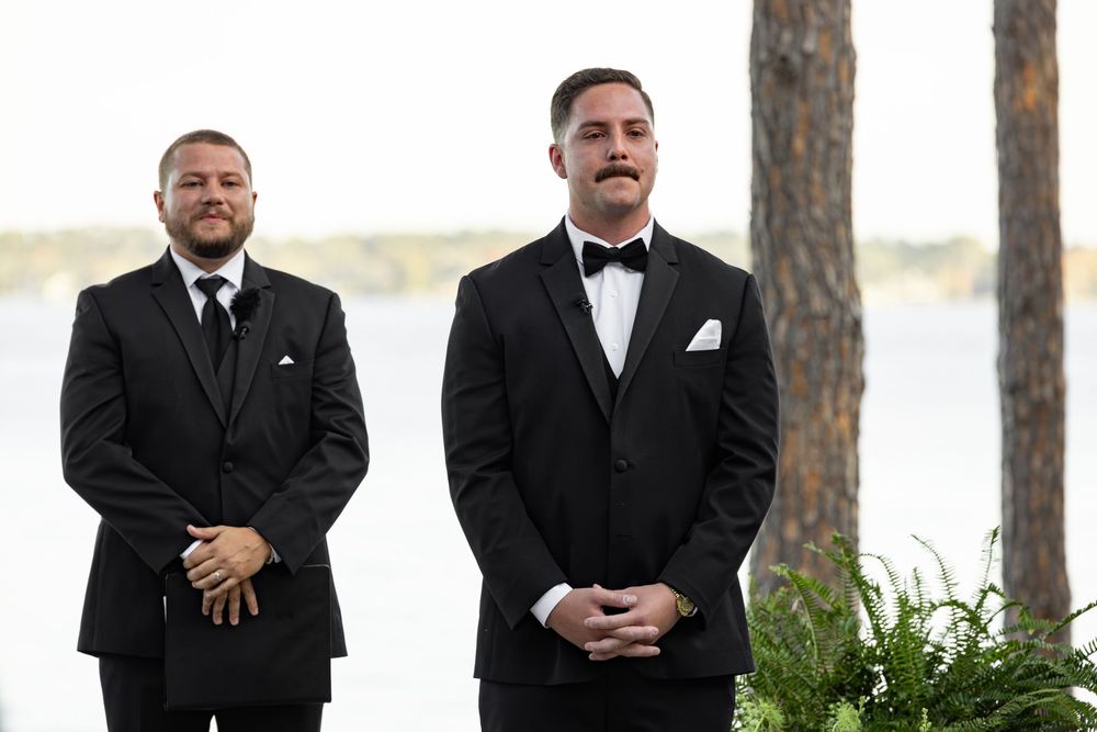 Two men in black tuxedos and bow ties stand together outdoors near pine trees during a formal event.