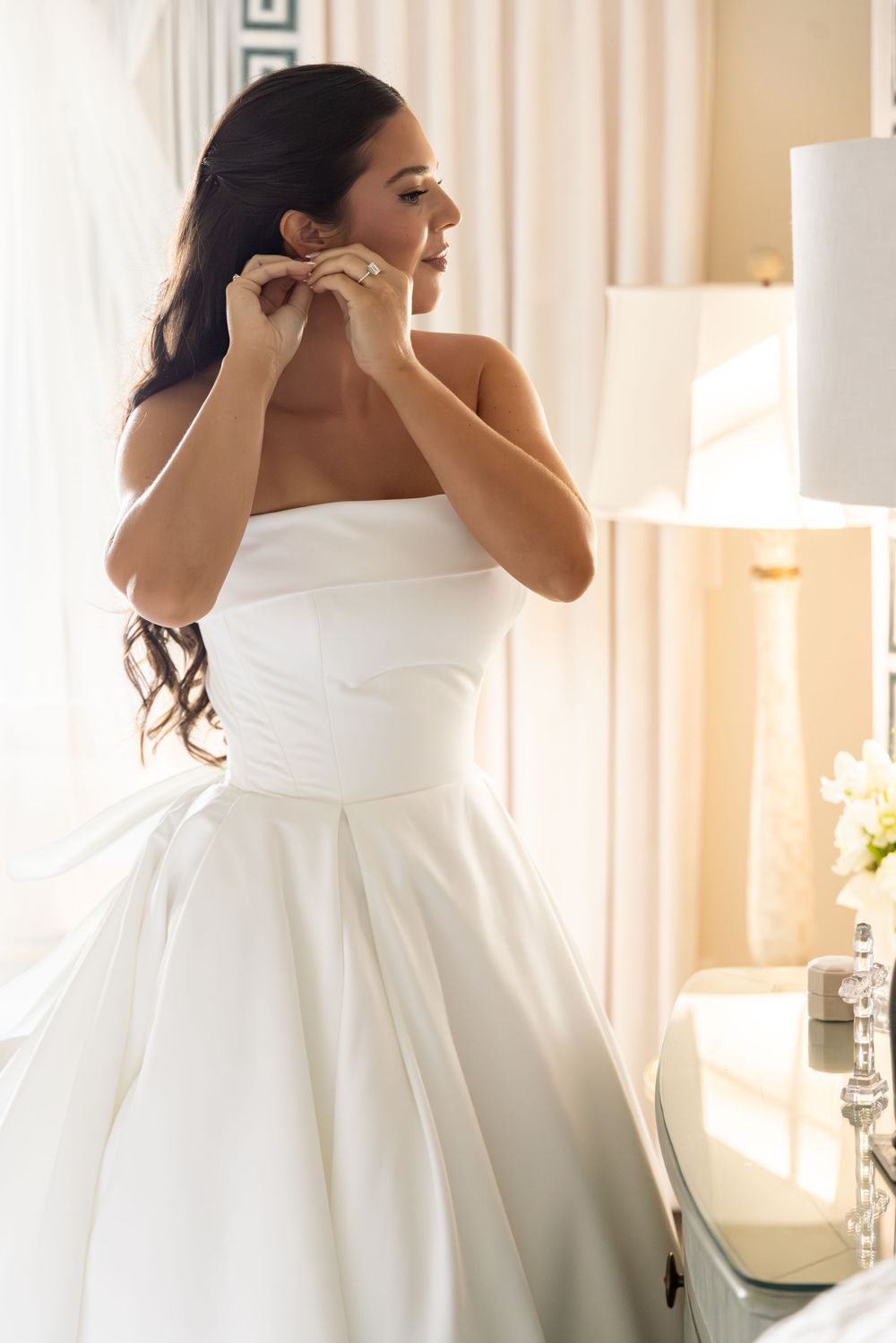 Bride putting on elegant pearl drop earrings while getting ready in a sunlit room with white curtains.