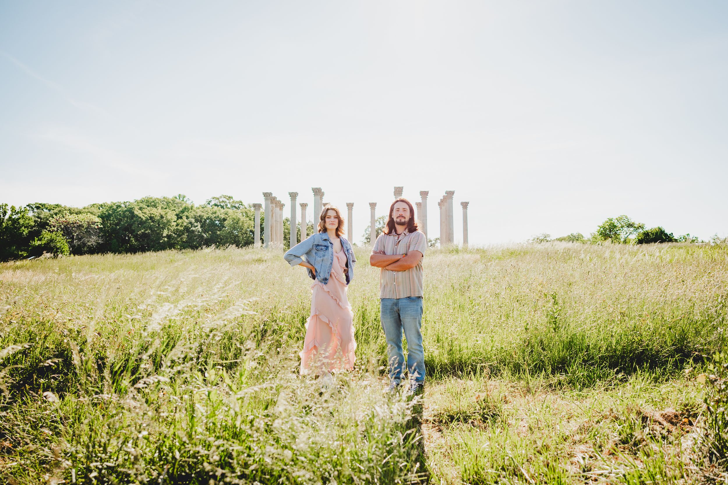 A couple walks through a sunny meadow filled with tall grass and wildflowers on a bright summer day.