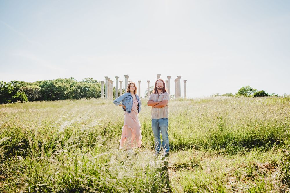 A couple walks through a sunny meadow filled with tall grass and wildflowers on a bright summer day.