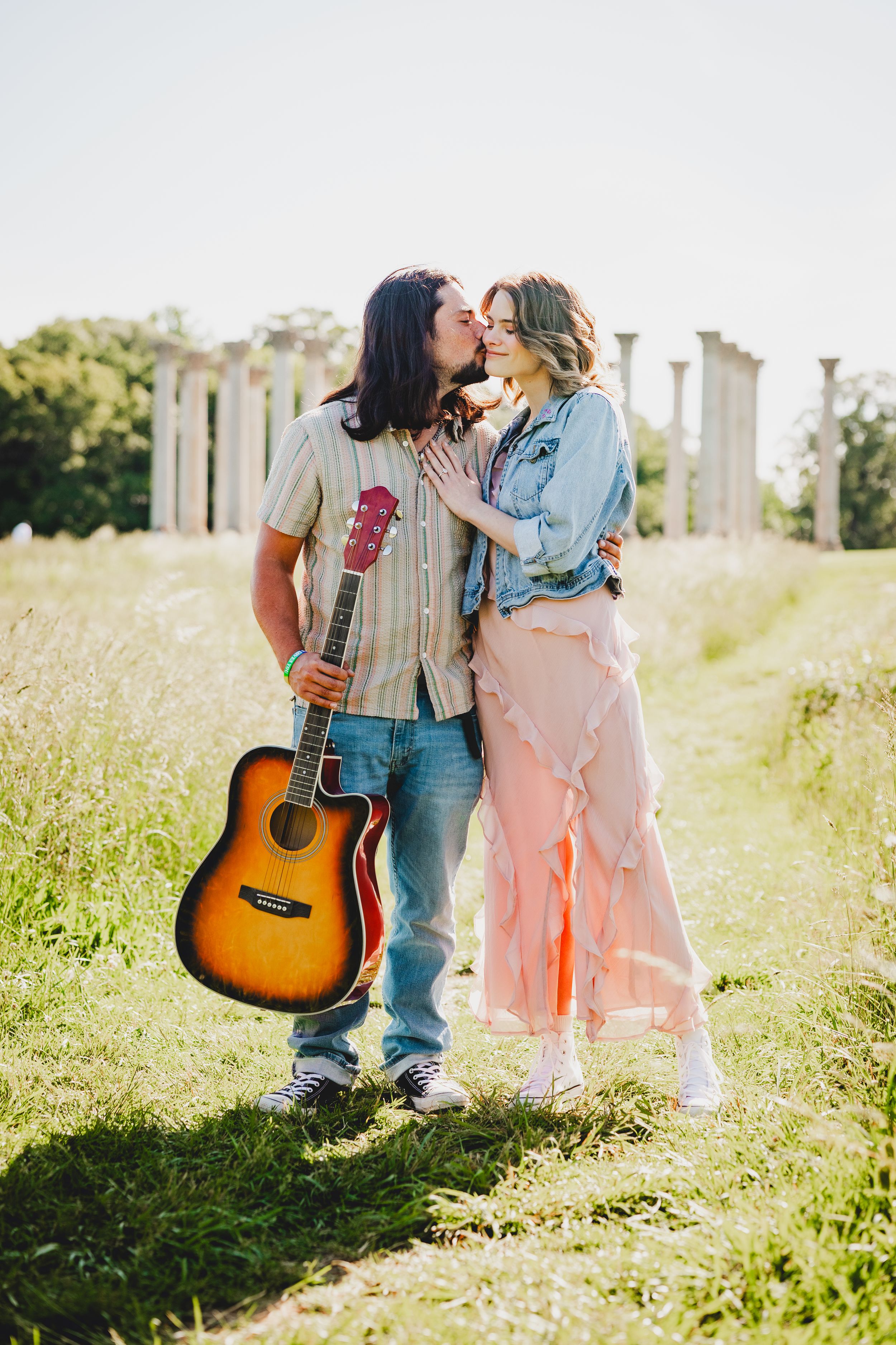 A couple stands in a sunlit meadow with a guitar, sharing a romantic moment in vintage-inspired outfits.