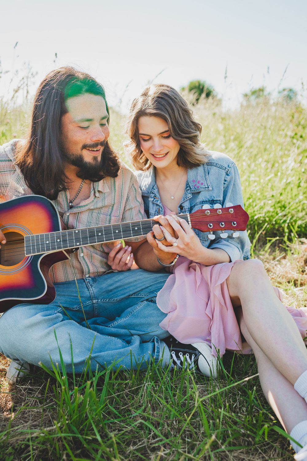 Couple relaxing in sunny meadow with acoustic guitar sharing intimate moments in casual denim and pink outfits.