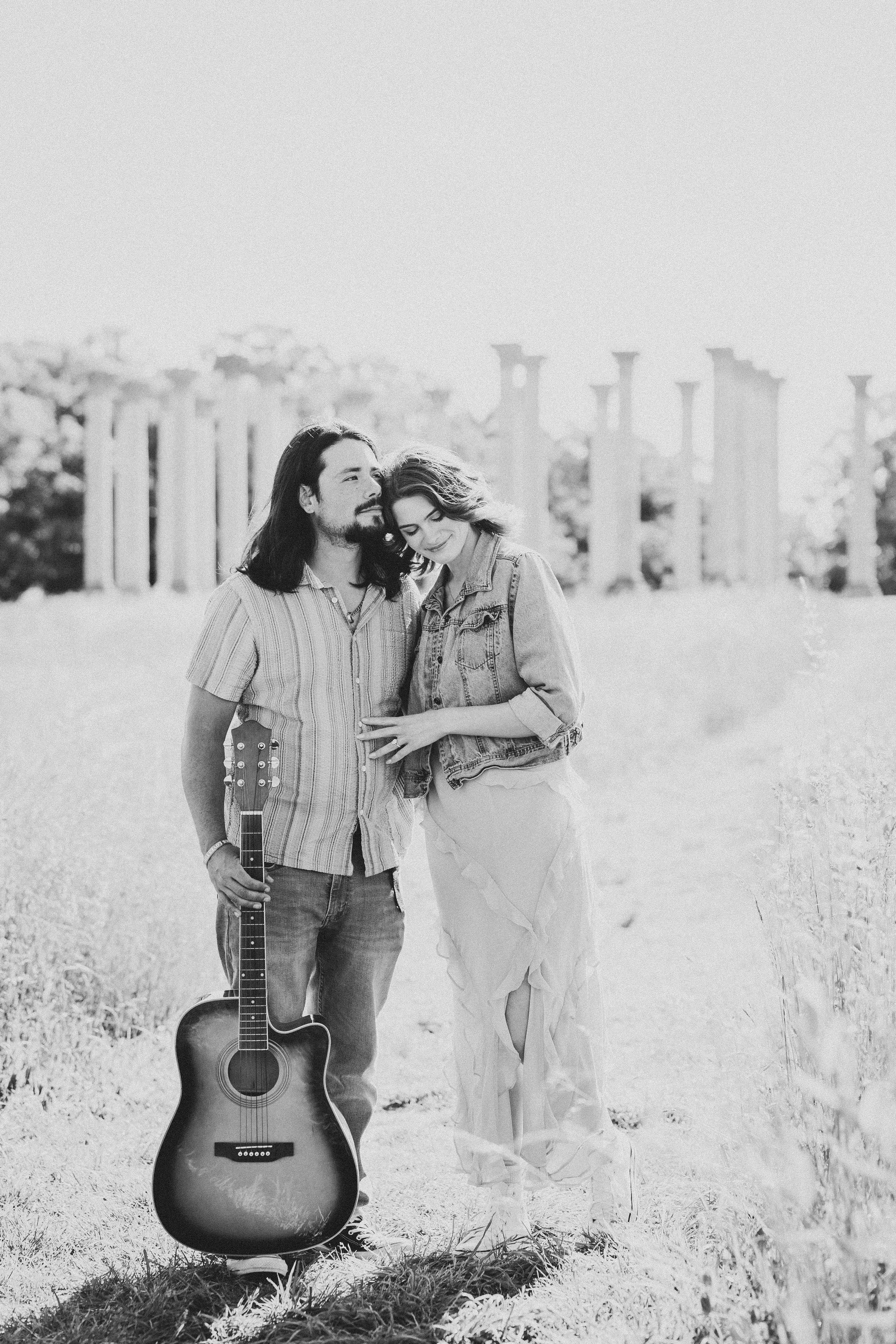 A couple shares a tender moment in a field while holding an acoustic guitar in black and white photography.