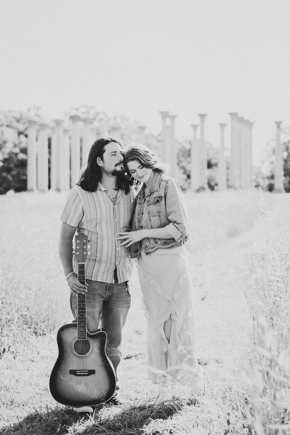 A couple shares a tender moment in a field while holding an acoustic guitar in black and white photography.