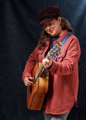 A musician in a pink sweatshirt and baseball cap plays an acoustic guitar against a dark blue backdrop.