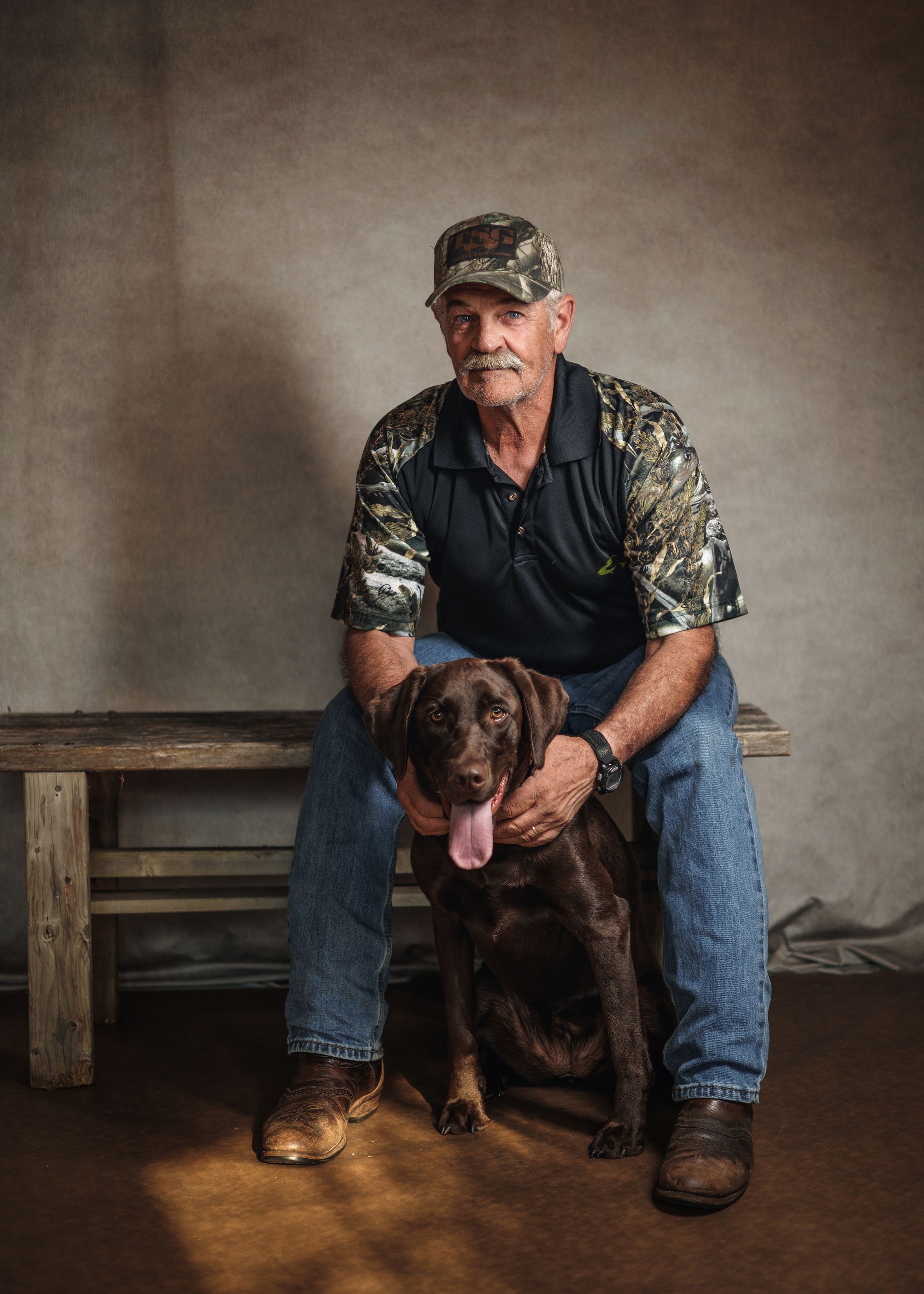 A chocolate Labrador retriever sits beside someone wearing jeans and camouflage in a rustic indoor setting.