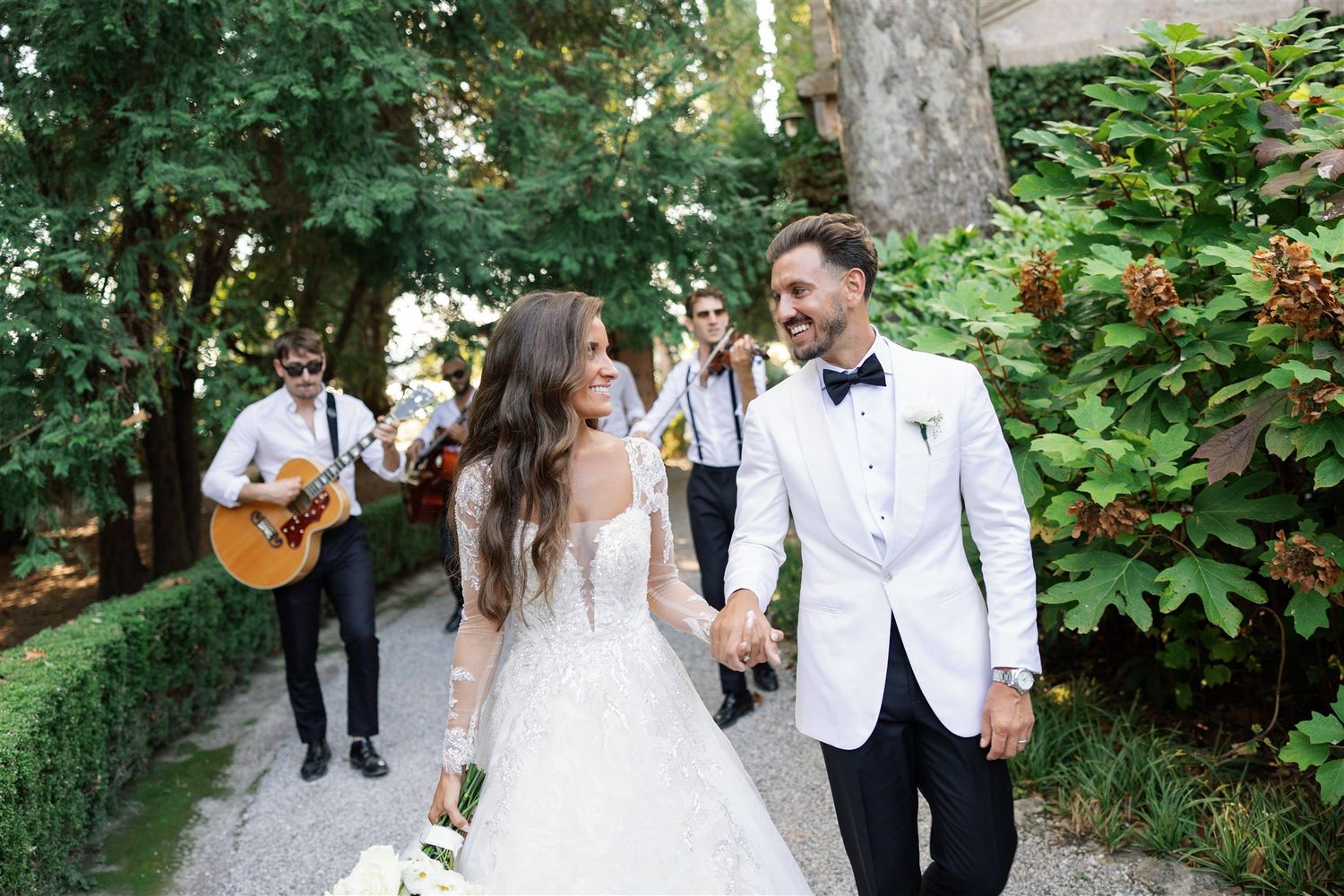 Bride and groom walking in the gardens of Isola del Garda accompanied by a roaming band
