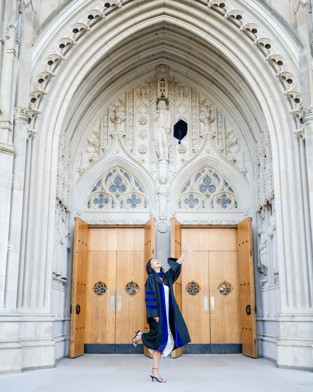 Person in a navy blue graduation gown jumps for joy in front of ornate white gothic cathedral arches.