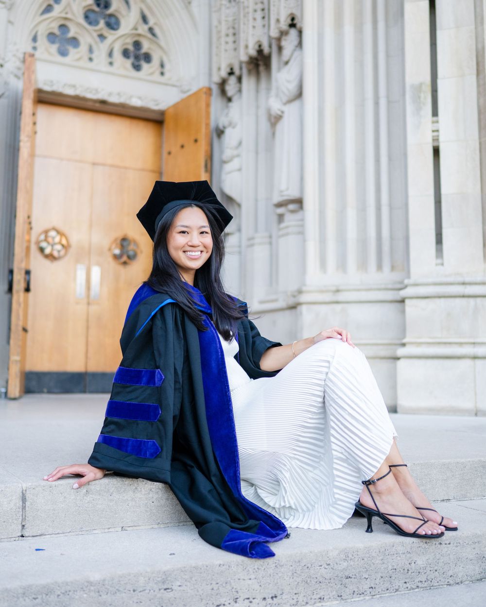 Graduate in cap and gown sits on stone steps of historic building for graduation photos.
