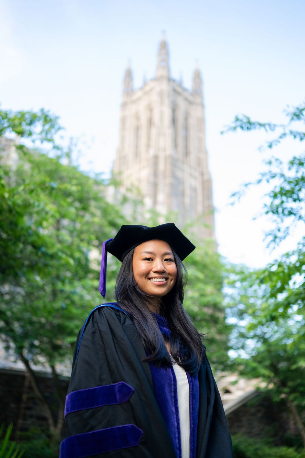 Graduate in academic regalia poses on a sunny day with a gothic university building visible in the background.