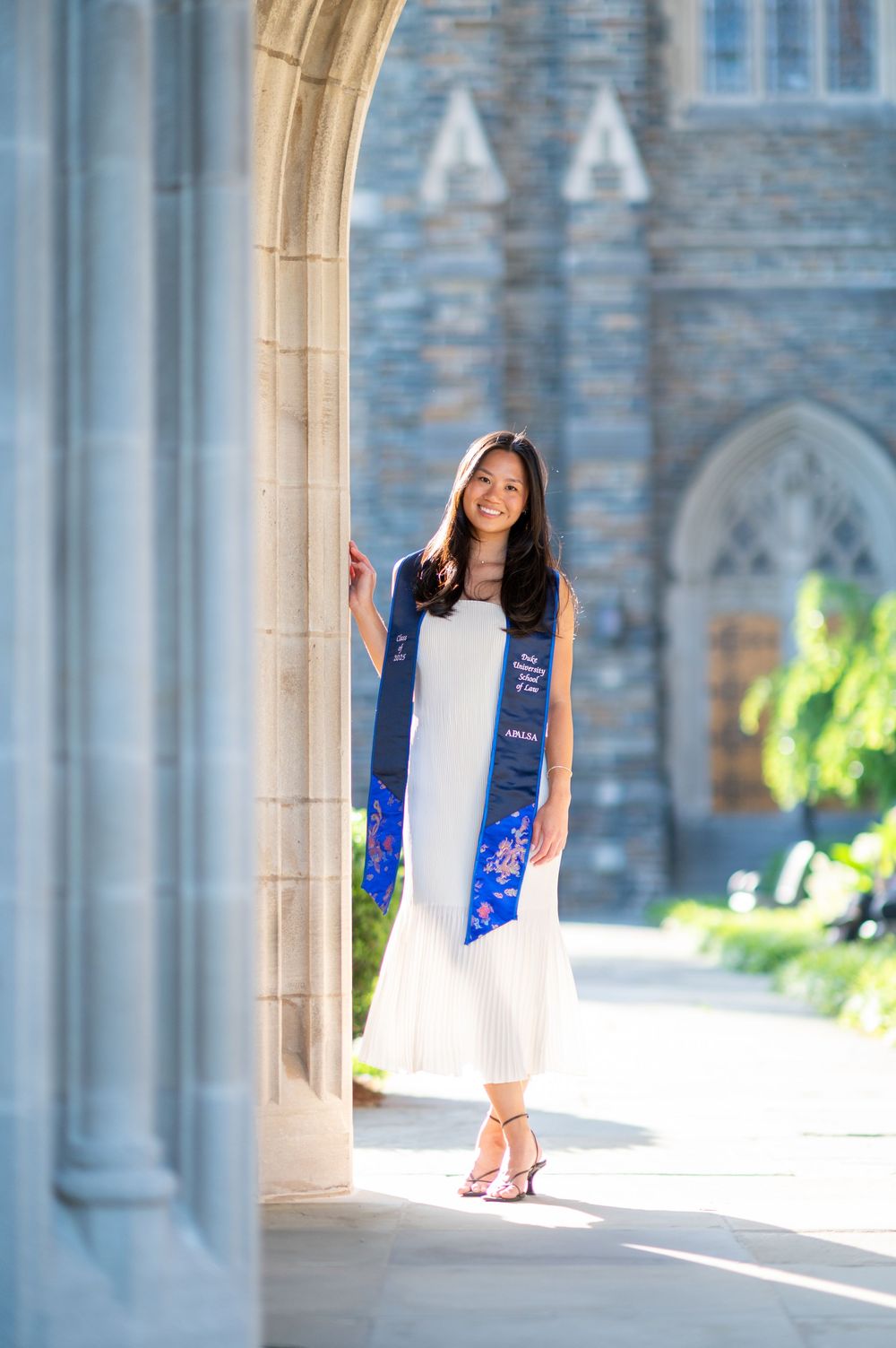 Graduate in white dress and blue stole poses in front of Gothic stone archways during graduation photoshoot.