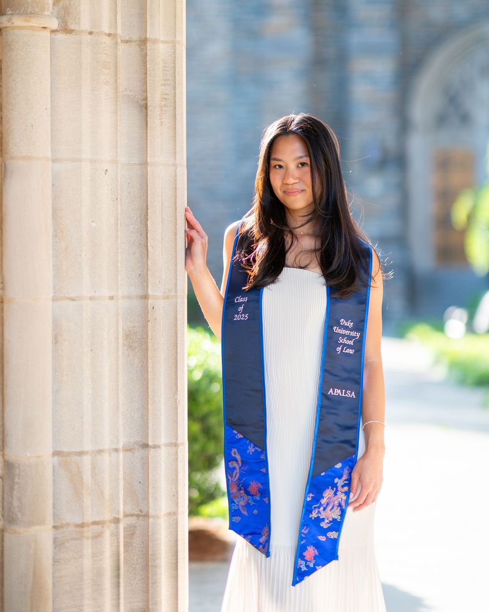 A graduate in a blue floral dress and navy sash poses elegantly against classical stone columns in outdoor portraits.