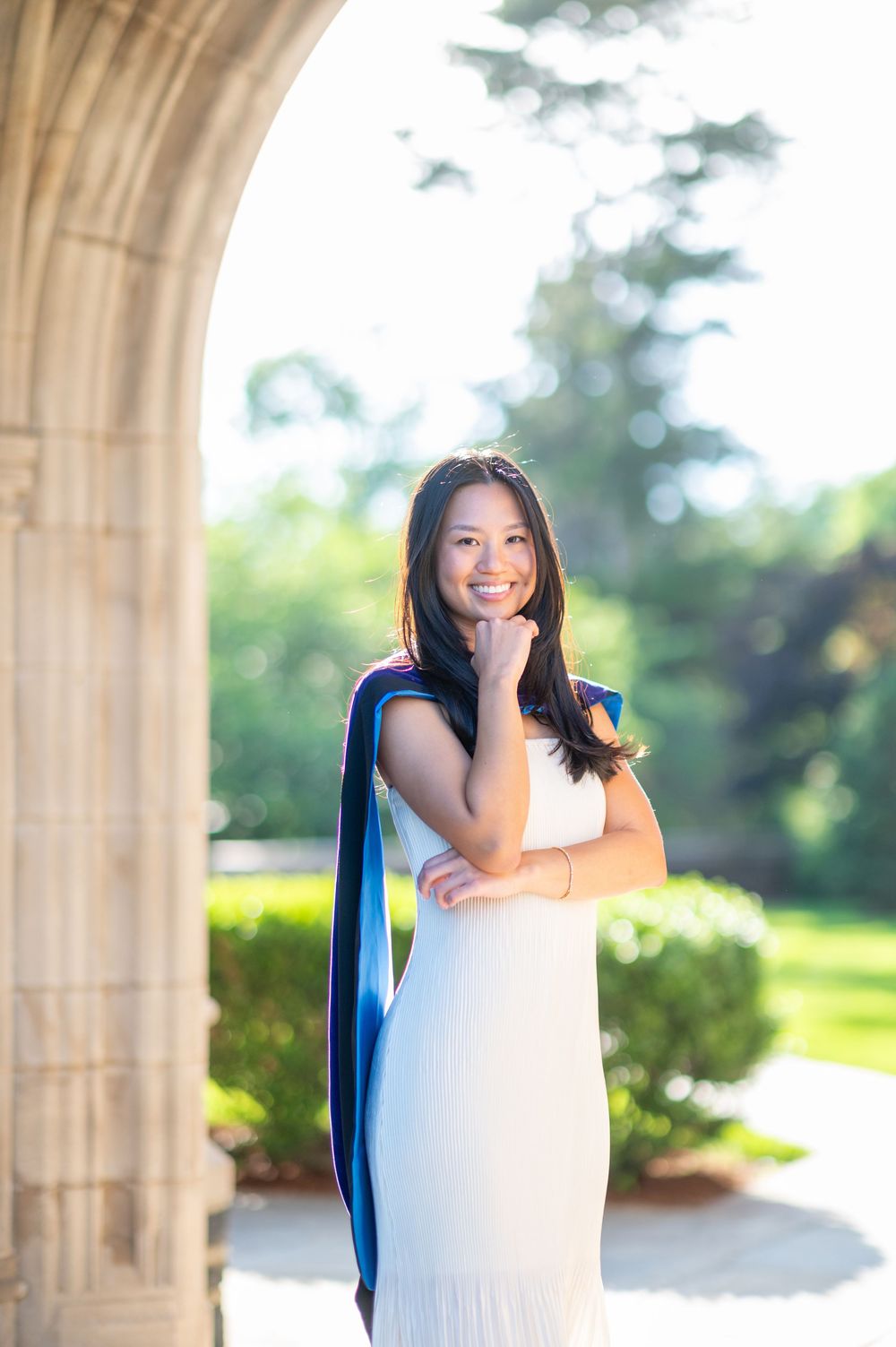 A person in a white dress poses gracefully under stone archways in an outdoor garden setting.
