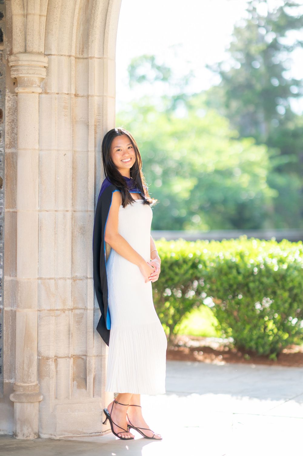 Graduate in white dress and cap poses under ornate stone archways on a sunny day.