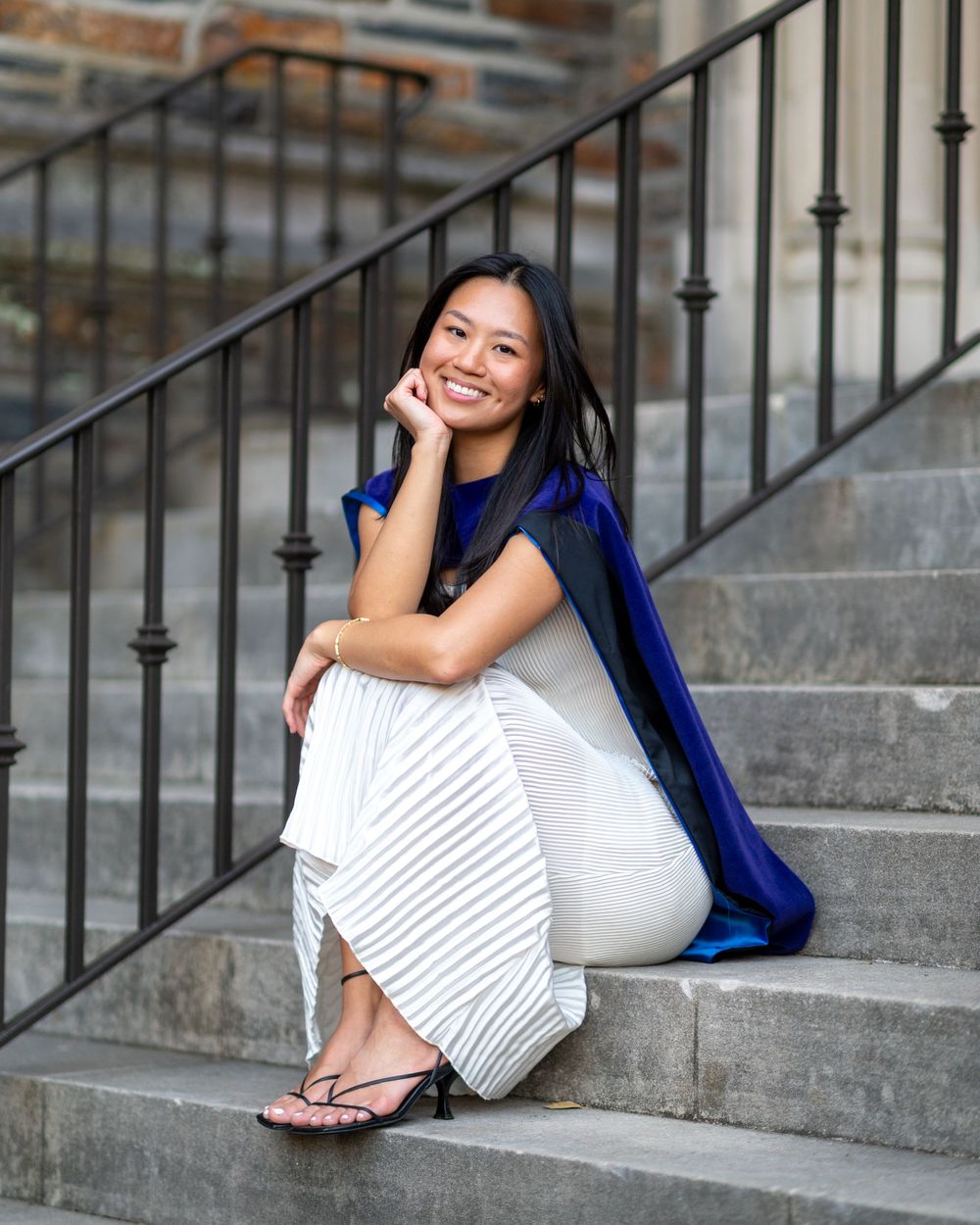 Someone in a white dress and blue blazer sits casually on concrete steps while smiling and leaning against a metal railing.
