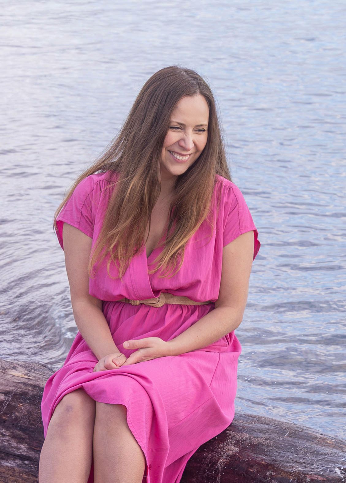 Person in bright pink dress sitting by the water's edge on a sunny day, smiling and looking away from camera.