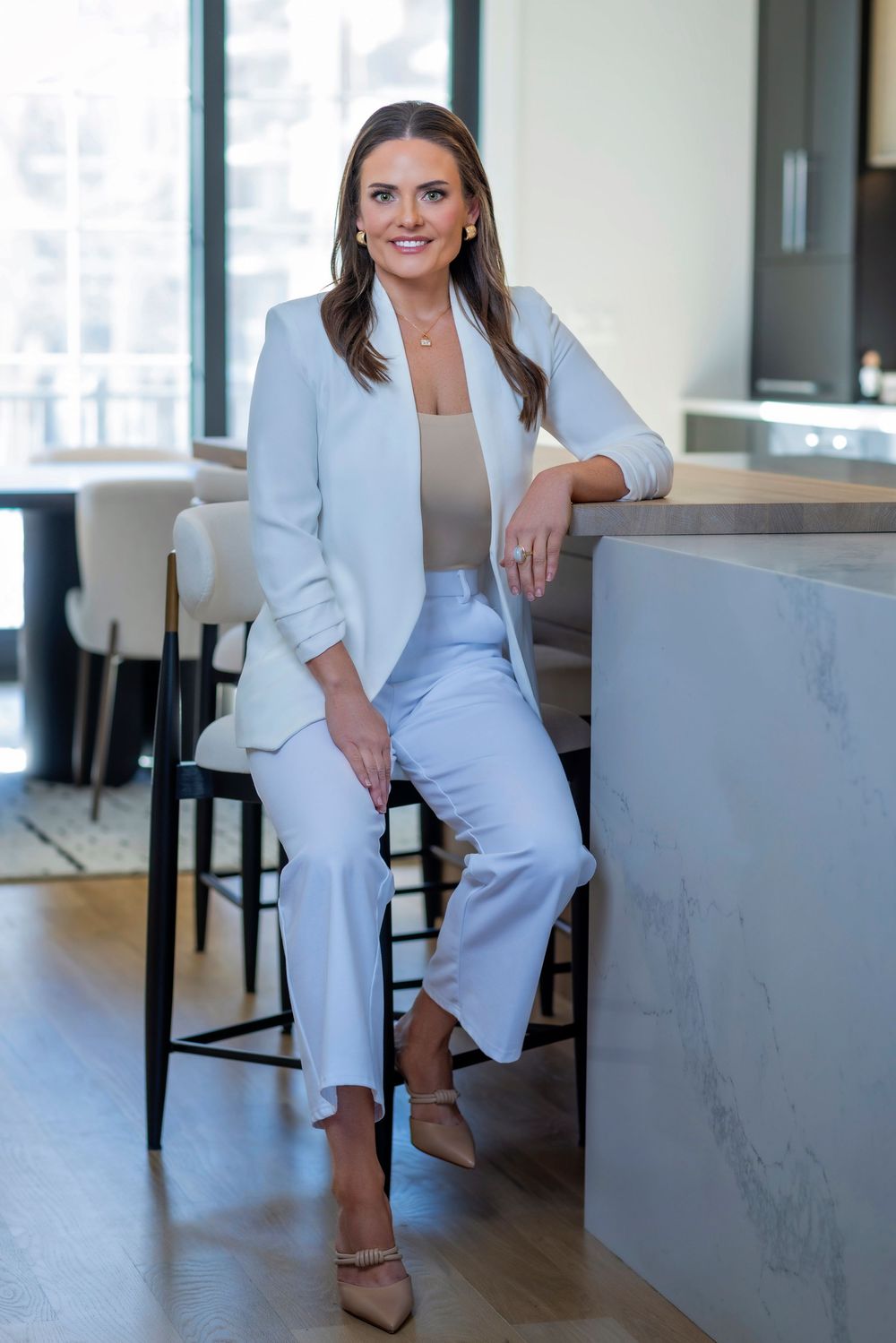 Professional wearing a crisp white blazer and pants with nude top sits casually at modern marble counter.