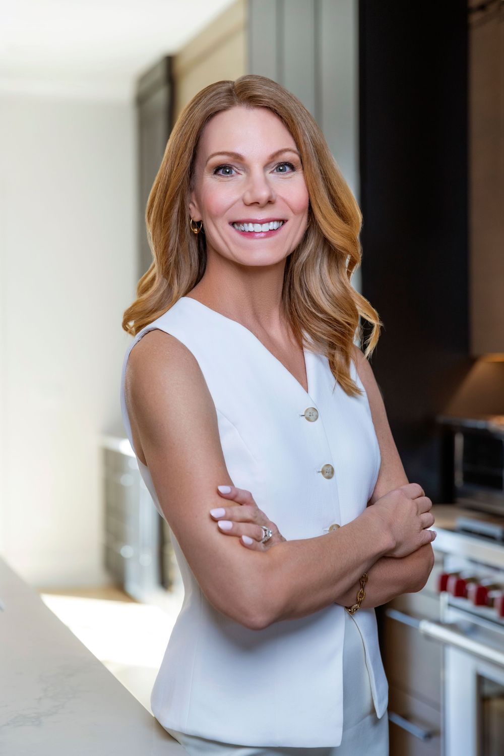 Professional portrait of a woman in a white sleeveless top smiling warmly in a modern kitchen setting.