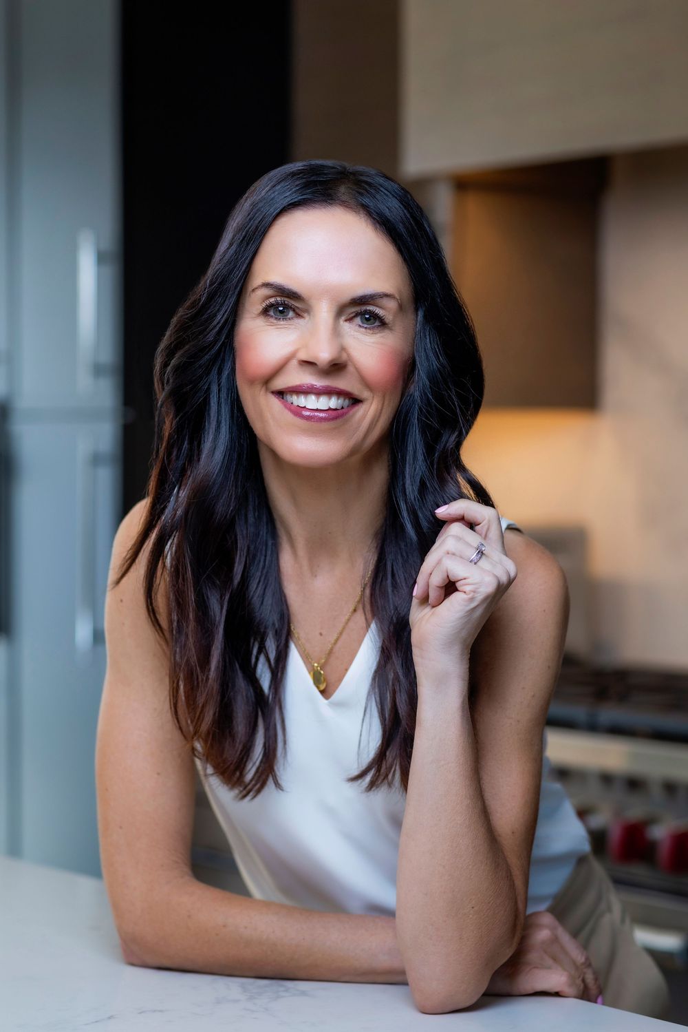 A smiling person with dark hair wearing a white sleeveless top leans on a surface in a well-lit interior setting.