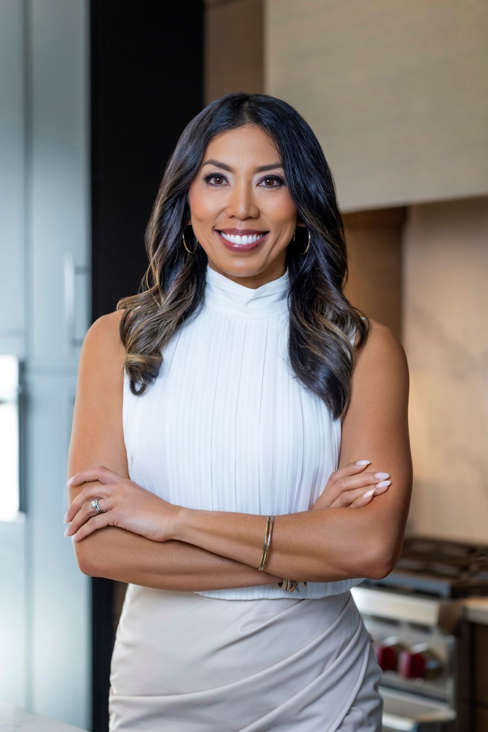 Professional portrait of someone in a white turtleneck top and gray skirt smiling warmly with arms crossed.