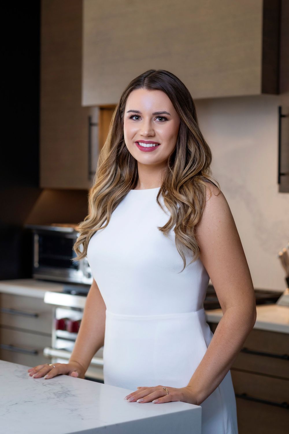 A person in a white sleeveless dress stands at a marble countertop in a modern kitchen interior.