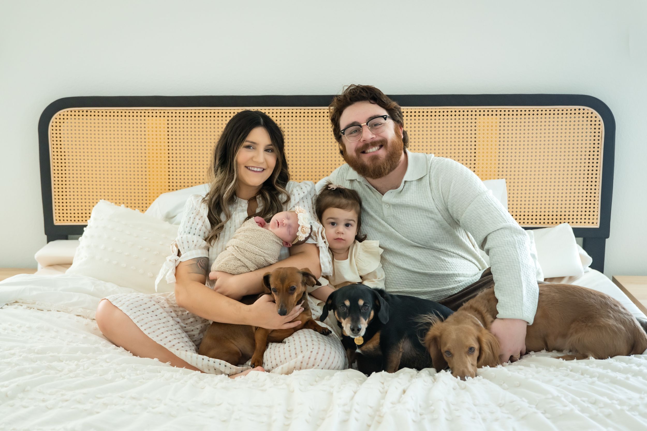 A family relaxes together on a white bed with their small dog while smiling at the camera.