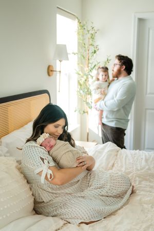 Family members share tender moments with newborn baby on white bedding in bright, airy bedroom.