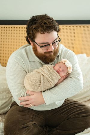 Parent wearing white sweater holds sleeping newborn baby wrapped in blanket while resting on couch.