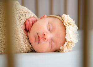 Sleeping baby wearing a peach flower headband rests peacefully against a cream-colored blanket.