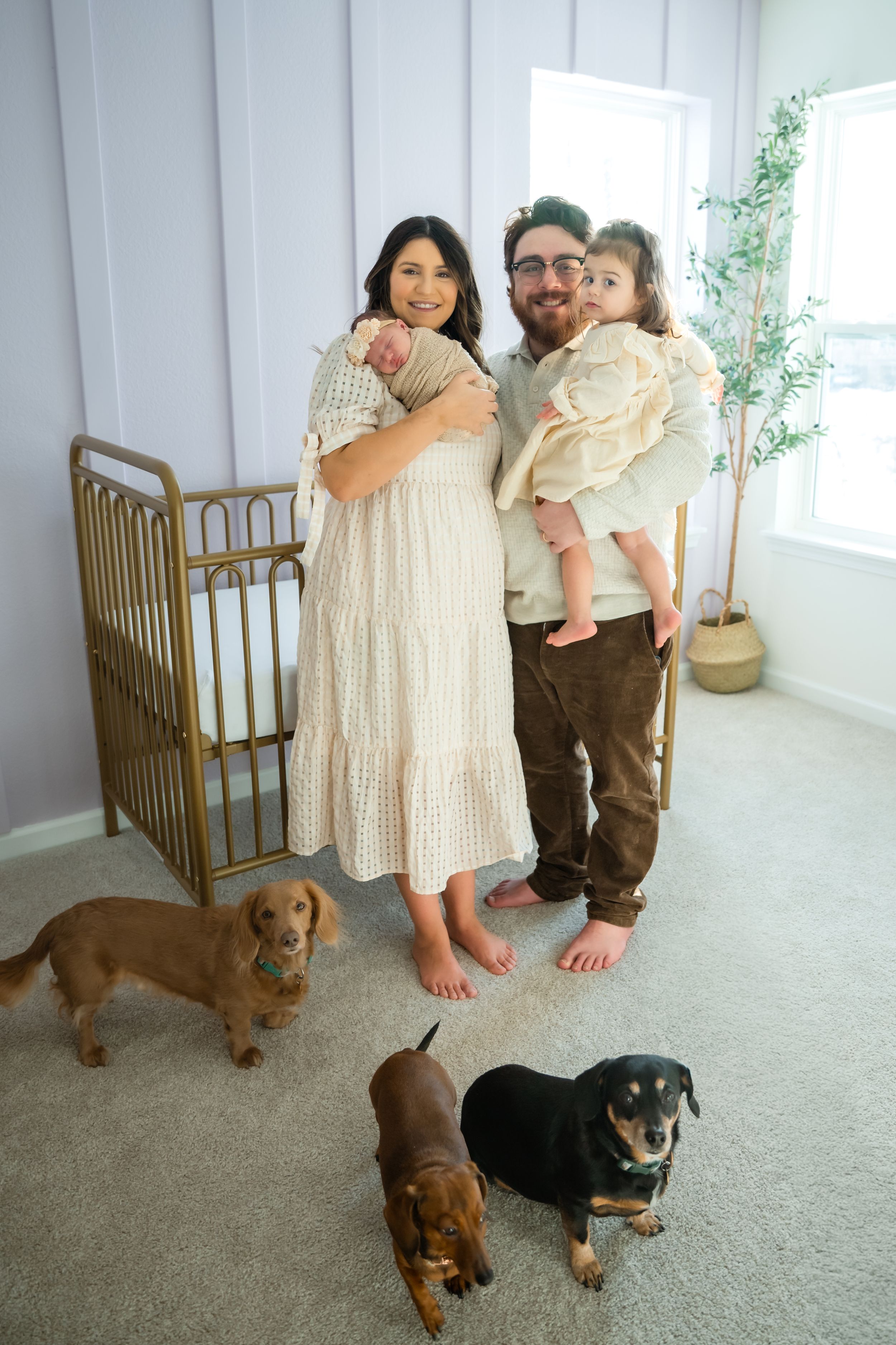 A couple poses with their dachshund dogs in a bright white room with a wooden crib.