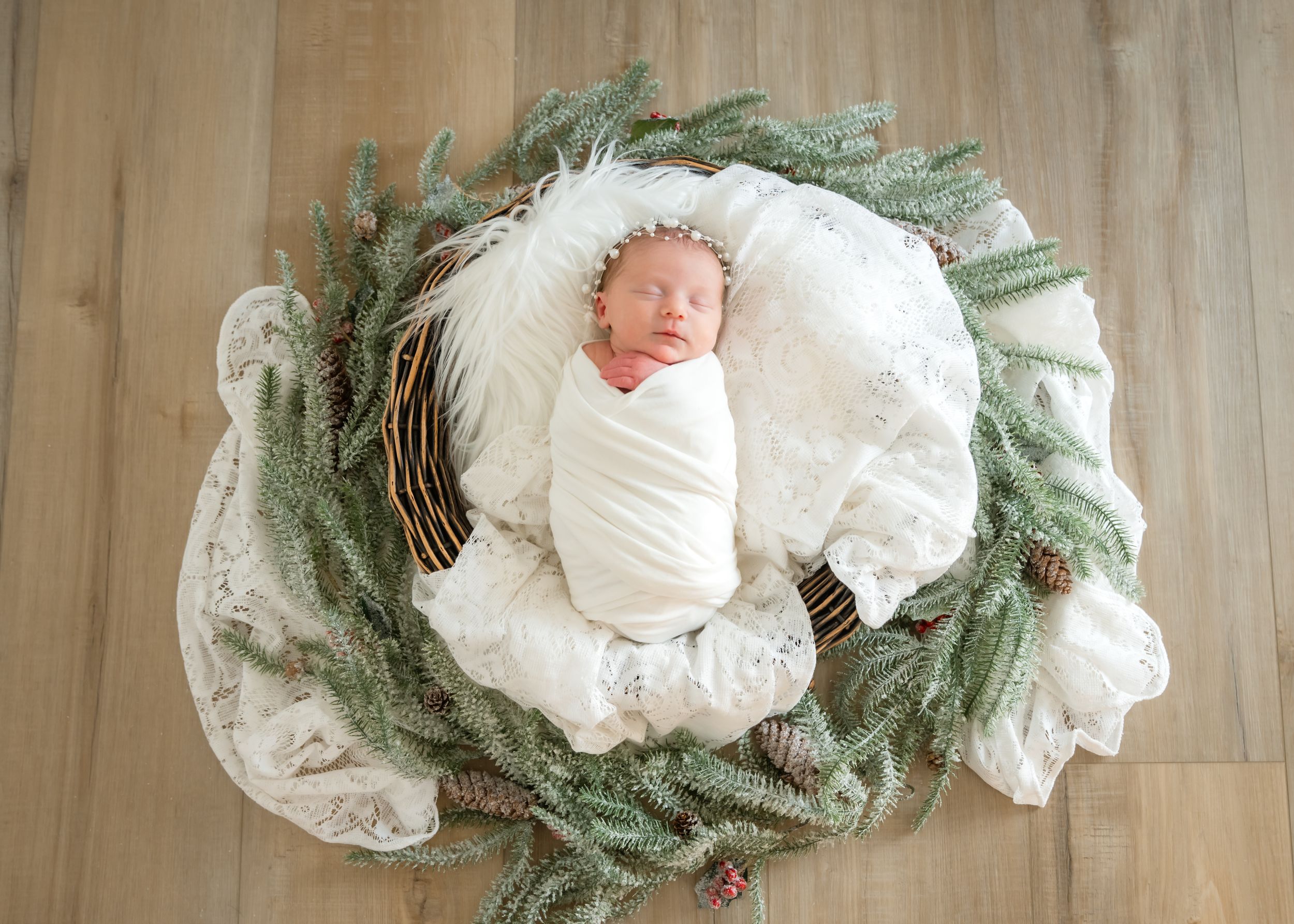 Newborn baby swaddled in white blanket sleeping in wicker basket surrounded by evergreen branches and white lace.