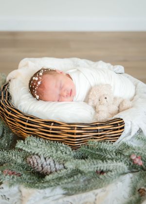 A sleeping newborn swaddled in white fabric rests peacefully in a woven basket on a bed of evergreen branches.