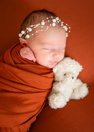 A sleeping newborn baby wrapped in a rust-colored blanket with small white flowers in their hair rests against a teddy bear.