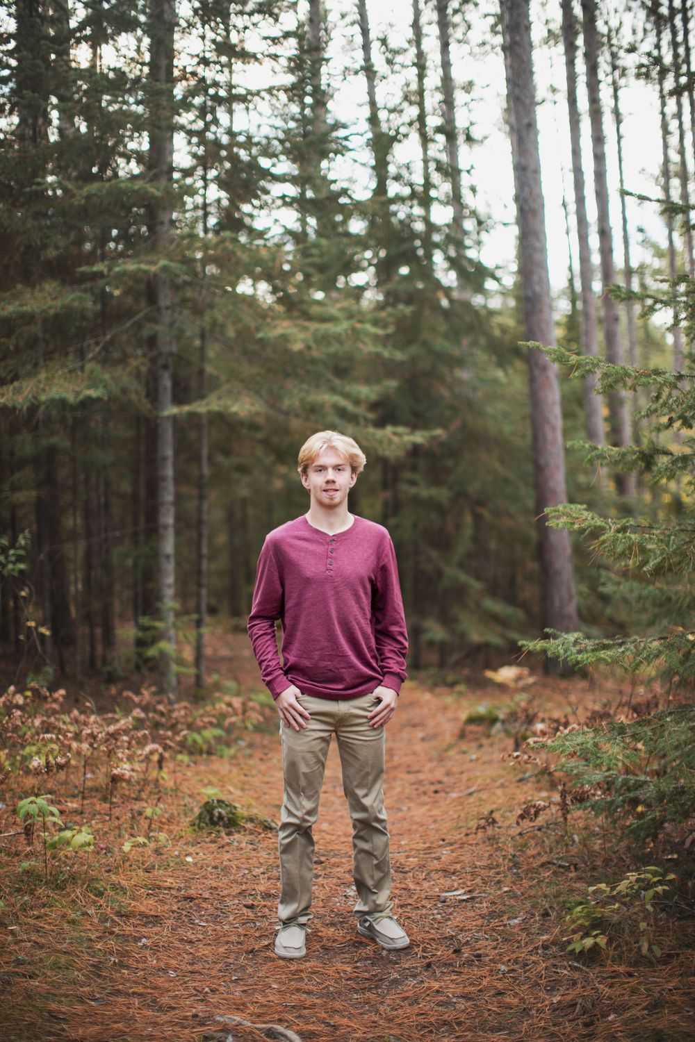 A person wearing a burgundy sweater and khaki pants stands on a forest trail surrounded by tall evergreen trees.