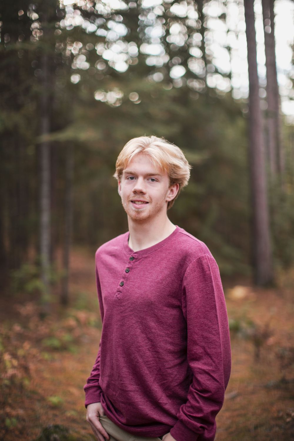 Portrait of someone wearing a burgundy sweater in a forest setting with blurred pine trees in the background.