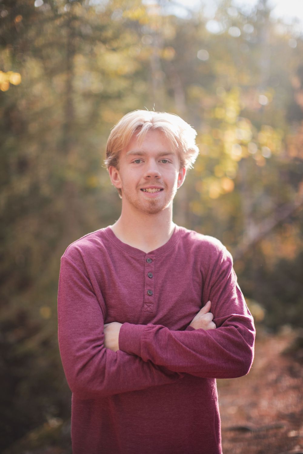 A person in a burgundy henley shirt poses with crossed arms in a sunny autumn forest setting.