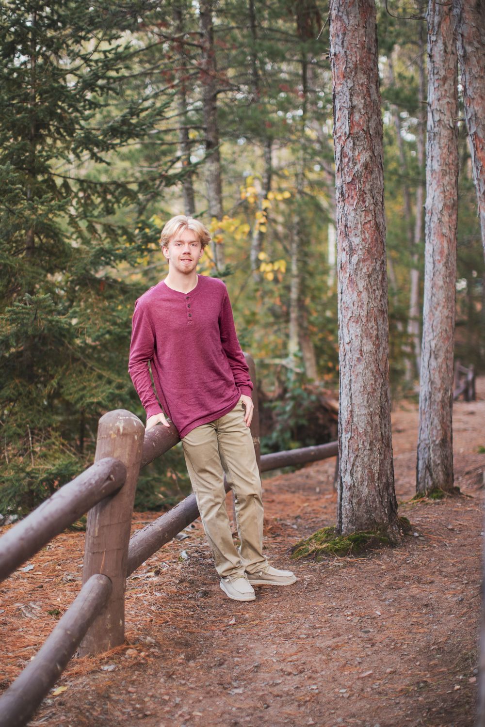Someone in a burgundy sweater and khaki pants stands casually by a wooden railing on a forest trail.