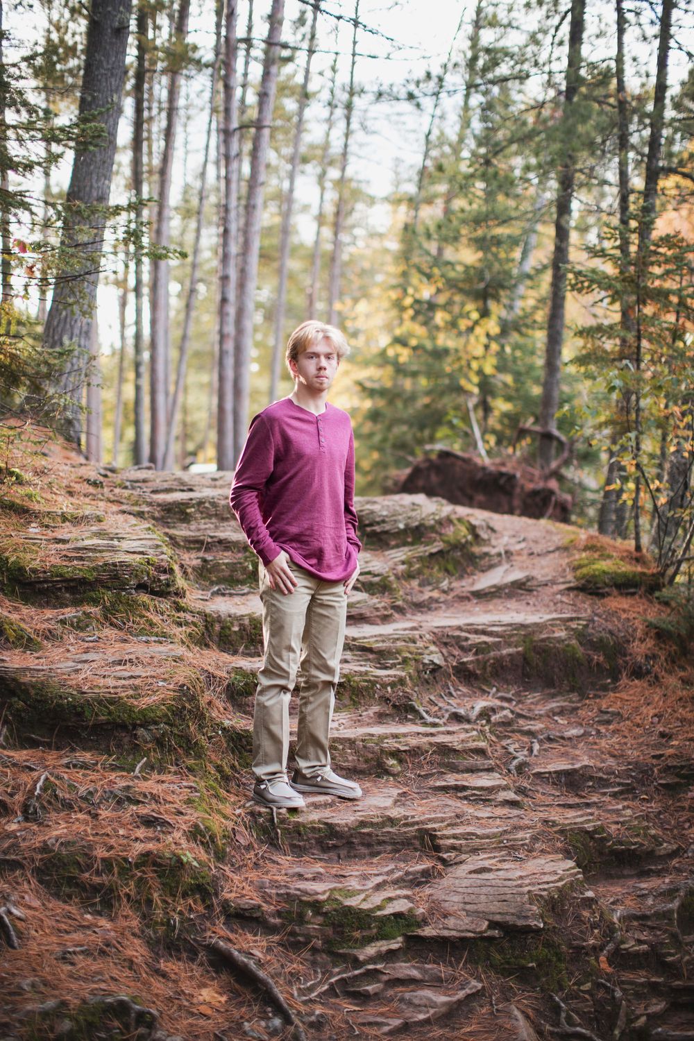 A person in a maroon sweater stands on a rocky forest path surrounded by tall pine trees.